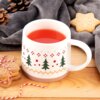 Na zdjęciu znajduje się porcelanowy kubek o pojemności 380 ml z motywem świątecznym, przedstawiającym zielone choinki i czerwone gwiazdki. Kubek stoi na drewnianej powierzchni, obok znajdują się ciasteczka, szyszki i szara tkanina.