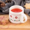 Na zdjęciu znajduje się porcelanowy kubek o pojemności 380 ml z czerwonym napojem, ozdobiony świątecznym wzorem z motywem gwiazdek i ostrokrzewu. Obok kubka leżą ciasteczka związane sznurkiem oraz szyszki, a w tle widoczny jest szary koc.