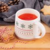 Na zdjęciu znajduje się porcelanowy kubek o pojemności 380 ml z czerwonym napojem, ozdobiony wzorami w kształcie śnieżynek, serc i fal. Obok kubka leżą ciasteczka związane sznurkiem oraz szyszki, a w tle widoczny jest szary koc.