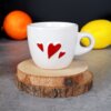 Biały ceramiczny kubek do espresso o pojemności 80 ml z trzema czerwonymi sercami na boku, ustawiony na drewnianej podstawce. W tle widoczne są pomarańczowy i żółty owoc.