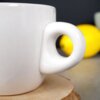 Zbliżenie na biały ceramiczny kubek do espresso o pojemności 80 ml z uchwytem w kształcie serca, ustawiony na drewnianej podstawce. W tle widoczny jest rozmyty żółty obiekt.