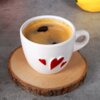 Biały ceramiczny kubek do espresso o pojemności 80 ml z czerwonymi sercami na boku, stoi na okrągłej drewnianej podstawce. W tle widoczny jest fragment żółtego owocu na szarej powierzchni.