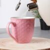 Różowy ceramiczny kubek z wytłoczonym wzorem serc stoi na drewnianej podstawce, a do jego wnętrza wlewana jest woda z czajnika. W tle widoczna jest rozmyta roślina w doniczce.
