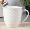 Biały ceramiczny kubek o pojemności 580 ml z wytłoczonym wzorem serc, ustawiony na drewnianej podstawce. W tle widoczny jest czajnik z drewnianym uchwytem oraz zielona roślina w doniczce.