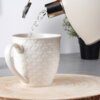 Biały ceramiczny kubek o pojemności 580 ml z wytłoczonym wzorem serc stoi na drewnianej podstawce, a do jego wnętrza nalewana jest woda z czajnika. W tle widoczna jest roślina w czarnym doniczce.
