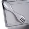 Szary pojemnik śniadaniowy z widocznym wewnątrz sztućcem łączącym funkcje łyżki i widelca. Pojemnik ma gładką powierzchnię i prostokątny kształt.