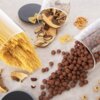 Na zdjęciu widoczne są trzy szklane pojemniki z pokrywkami, z których wysypują się różne produkty sypkie: makaron spaghetti, suszone grzyby oraz kulki zbożowe. Obok znajdują się również pojedyncze kawałki makaronu kokardki, precelki, suszone grzyby i ziarna fasoli.