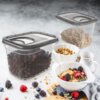 Na zdjęciu widoczne są dwa przezroczyste pojemniki z szarymi pokrywkami, wypełnione sypkimi produktami spożywczymi, obok których znajdują się miski z płatkami śniadaniowymi i owocami oraz mały dzbanek. Na blacie leżą rozsypane owoce i płatki, tworząc estetyczną kompozycję.