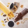 Na zdjęciu widoczne są trzy szklane pojemniki z pokrywkami, wypełnione makaronem spaghetti, suszonymi grzybami i kulkami czekoladowymi, które są rozsypane na blacie. Obok znajdują się różne produkty sypkie, takie jak makaron kokardki, suszone owoce, fasola, precelki i płatki śniadaniowe.