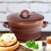 Na zdjęciu znajduje się brązowy, ceramiczny garnek z pokrywką i uchwytami po bokach, ustawiony na drewnianym blacie. Obok garnka leży kromka chleba z białą pastą i zielonymi dodatkami, a także gałązka natki pietruszki.