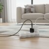 Na zdjęciu widać ładowarkę sieciową FORCELL F-Energy A130101-E 30W w kolorze czarnym, podłączoną do białej listwy zasilającej, leżącej na dywanie z geometrycznym wzorem. W tle znajduje się jasna sofa z poduszkami, a pomieszczenie jest dobrze oświetlone naturalnym światłem wpadającym przez duże okno.