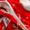 Zbliżenie na czerwony materiał z białym zamkiem błyskawicznym, ozdobiony białymi płatkami śniegu i elementami w kolorze białym, pomarańczowym i brązowym. W tle widoczne są fragmenty wzoru przypominającego świąteczne motywy.