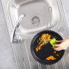 Na zdjęciu widoczny jest zlew z metalową baterią oraz patelnia stalowa GRANDE 26 cm, która jest czyszczona zieloną gąbką. Wewnątrz patelni znajdują się resztki pomarańczowego jedzenia.