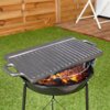 Dwustronna żeliwna płyta do grillowania o wymiarach 45,4x23 cm umieszczona na okrągłym grillu z płonącym węglem, w tle drewniana ściana i zielona trawa. Płyta ma uchwyty po obu stronach i widoczne rowki na powierzchni.