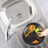 Na zdjęciu widać stalową patelnię COOKCELL 24 cm, która jest czyszczona zieloną gąbką nad zlewem. Na powierzchni patelni znajdują się resztki jedzenia w pomarańczowym kolorze.