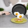 Na zdjęciu widać granitową patelnię nieprzywierającą, z której zsuwają się dwa jajka sadzone na żółty talerz z tostami. W tle znajdują się rękawica kuchenna, rośliny doniczkowe i fragmenty kuchennego blatu.