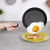Na zdjęciu widoczna jest ręka trzymająca granitową patelnię, z której zsuwane jest jajko sadzone na talerz z tostami. Talerz jest żółty, a w tle znajdują się elementy kuchenne, takie jak roślina i drewniane akcesoria.