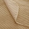 Beżowa narzuta z weluru o wzorze jodełki, z widocznym zagięciem ukazującym wewnętrzną stronę materiału. Powierzchnia narzuty jest pikowana w regularne, geometryczne wzory.