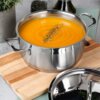Stalowy garnek z pokrywką o pojemności 3 litrów i średnicy 20 cm, wypełniony pomarańczową zupą, stoi na drewnianej desce. Obok garnka znajduje się pokrywka, a w tle widoczne są rośliny i butelka z oliwą.