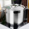 Stalowy garnek z pokrywką o pojemności 6,5 litra i średnicy 22 cm stoi na kuchence, otoczony różnymi kuchennymi akcesoriami i pojemnikami z produktami spożywczymi. Garnek ma błyszczącą powierzchnię i dwa uchwyty po bokach.