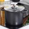 Czarny garnek granitowy z dwoma uchwytami i szklaną pokrywką z metalowym uchwytem, stoi na kuchence. W górnym rogu widoczne są ikony z napisami: "INDUCTION", "GAS", "ELECTRIC", "CERAMIC".