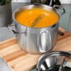 Na zdjęciu widoczny jest stalowy garnek z pomarańczową zupą, posypaną przyprawami, stojący na drewnianej desce. Obok garnka leży pokrywka.