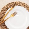 Na zdjęciu znajduje się biały, porcelanowy talerz obiadowy o falistym brzegu, umieszczony na plecionym podkładce. Obok talerza leżą złoty widelec i nóż.