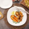 Na zdjęciu znajduje się talerz obiadowy z kawałkiem grillowanej ryby, pieczonymi ziemniakami, gałązkami rozmarynu i plasterkami cytryny. Obok talerza leżą sztućce, a w tle widoczne są winogrona i kwiaty.