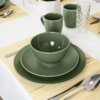 Na zdjęciu znajduje się zastawa stołowa w kolorze zielonym, w tym talerz deserowy ceramiczny o średnicy 21,5 cm, miska, kubek oraz sztućce na bambusowej podkładce. W tle widoczna jest szklanka z wzorem oraz biały obrus.