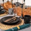 Na zdjęciu widać elegancko nakryty stół z czarnymi ceramicznymi talerzami deserowymi, sztućcami w kolorze miedzianym oraz czarnymi kubkami. Na stole znajdują się również szklanki, świeczniki i dekoracje z tkaniny w odcieniach brązu oraz zielone serwetki z ozdobnymi pierścieniami.