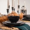 Na zdjęciu widoczny jest elegancko nakryty stół z czarną miseczką ustawioną na szarym talerzu deserowym ceramicznym, otoczonym przez czarne świeczniki z zapalonymi świecami. W tle znajdują się dekoracje z pomarańczowego materiału oraz zielona serwetka z białym wzorem.