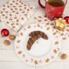 Na zdjęciu znajduje się porcelanowy talerz deserowy o średnicy 20 cm, ozdobiony wzorami pierników, serc i lasek cukrowych, na którym leżą dwa ciastka polane czekoladą. Obok talerza widoczne są czerwony kubek z napojem, serwetki z motywem pierników, orzechy włoskie, czerwona bombka oraz stos ciastek związanych sznurkiem.