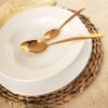 Na zdjęciu znajduje się biały, porcelanowy talerz obiadowy o średnicy 27 cm, na którym leżą dwie złote łyżki. Talerz jest umieszczony na plecionym podkładzie, a obok widoczna jest jasna serwetka.