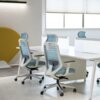 Na zdjęciu znajduje się nowoczesne biuro z białymi biurkami i ergonomicznymi krzesłami biurowymi w kolorze niebieskim, wykonanymi z tkaniny i siatki. W tle widoczne są duże okna z roletami oraz żółty element dekoracyjny.