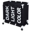Czarny, trzykomorowy kosz na pranie na kółkach z białymi napisami "DARK", "LIGHT" i "COLOR" na każdej z komór. Konstrukcja kosza jest wsparta na białych rurkach i wyposażona w cztery kółka.