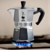 Srebrno-czarna kawiarka Bialetti Moka Express o pojemności 100 ml stoi na palniku gazowym z niebieskim płomieniem. Na kawiarkę widoczne są napisy: "Bialetti", "Moka Express", "Made in Italy".