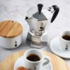 Srebrno-czarna kawiarka z logo Bialetti i napisem "Moka Express Made in Italy" stoi na blacie obok białej cukiernicy z drewnianą pokrywką oraz dwóch filiżanek z kawą na spodkach, z widocznym logo producenta. Na drewnianej podstawce leżą dwa ciastka.