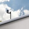 Na zdjęciu widoczny jest wiatromierz zamontowany na dachu budynku, na tle nieba z białymi chmurami. Urządzenie ma cylindryczny kształt i jest przymocowane do pionowego wspornika.