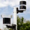 Na zdjęciu widoczny jest wiatromierz zamontowany na białym słupku, z dwoma czarnymi cylindrycznymi elementami. Na jednym z elementów znajduje się logo "Netatmo", a w tle widać rozmyte drzewa i niebo.