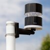 Na zdjęciu widoczny jest wiatromierz zamontowany na białym słupku, z czarną i białą obudową, na której znajduje się napis "Netatmo". W tle widać niebo z lekkim zachmurzeniem oraz fragment zieleni.