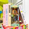 Miniaturowy domek Book Nook - Kocie miasteczko umieszczony pomiędzy kolorowymi książkami z widocznymi tytułami: 'magic girl', 'Vanilla ice cream', 'Street Fighter', 'PONY BROWN'.
