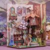 Miniaturowy domek Book Nook - Koncert leśnych kwiatów przedstawia szczegółową scenę z elementami roślinnymi, drewnianymi konstrukcjami i dekoracjami w stylu leśnym. W tle widoczne są ilustracje drzew i kwiatów, a na pierwszym planie znajdują się miniaturowe meble i ozdoby.