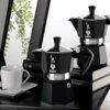 Czarne kawiarki Bialetti Moka Express stoją na stosie książek, obok białej filiżanki na spodku z łyżeczką. Na kawiarkach widoczne jest logo Bialetti oraz napis "Moka Express".