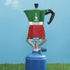 Zielono-czerwona kawiarka z logo "Bialetti" i napisem "Moka Express" umieszczona jest na metalowym palniku, który stoi na niebieskiej butli gazowej. W tle widać niebieskie niebo i zieloną trawę.