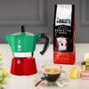 Zielono-czerwona kawiarka z logo "Bialetti" stoi obok opakowania kawy "Bialetti Perfetto Moka Classico Intensità 7", a na stole znajduje się filiżanka espresso i kilka kolorowych makaroników.