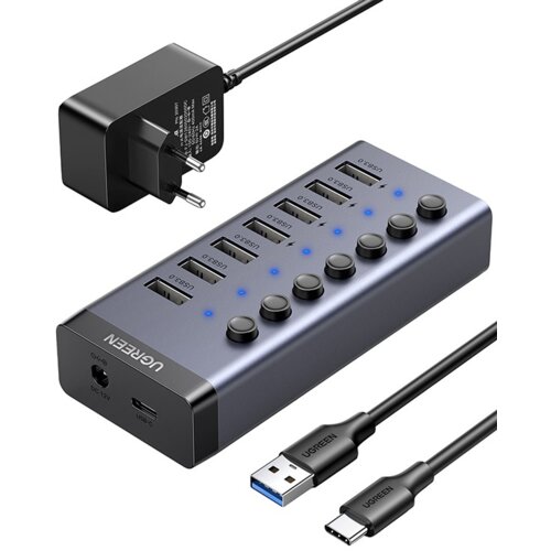 Na zdjęciu znajduje się hub USB marki UGREEN z siedmioma portami USB oznaczonymi jako "USB3.0" oraz przyciskami i niebieskimi diodami. Obok huba widoczny jest zasilacz z wtyczką oraz dwa kable USB, z których jeden ma końcówkę USB Typu C, a drugi USB Typu 