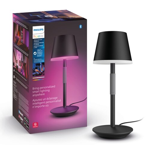 Na zdjęciu znajduje się czarna lampa stołowa obok pudełka z napisem "PHILIPS hue white and color ambiance" oraz tekstem "Bring personalized smart lighting anywhere". Pudełko jest ozdobione grafiką przedstawiającą lampę emitującą różowe światło.