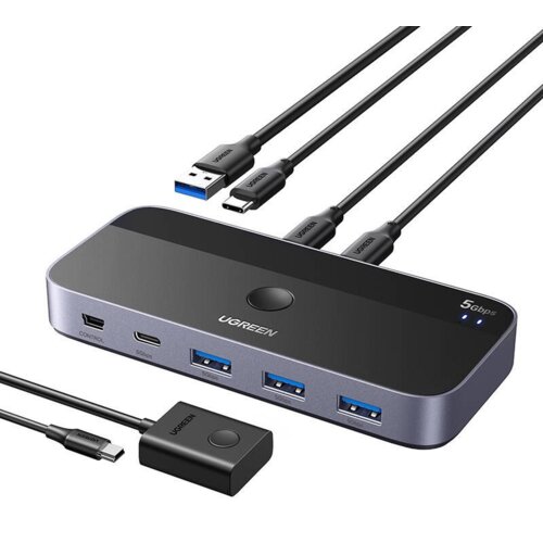 Na zdjęciu znajduje się przełącznik KVM marki UGREEN z kilkoma portami USB i kablami, z widocznym napisem "UGREEN" oraz oznaczeniem "5Gbps". Produkt ma elegancki, nowoczesny design z czarną i srebrną kolorystyką.