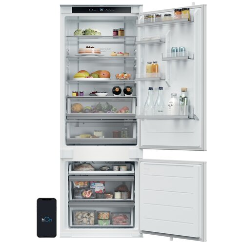 Otwarta lodówka z widocznymi półkami wypełnionymi różnorodnymi produktami spożywczymi, takimi jak owoce, warzywa, napoje i słoiki. Na drzwiach lodówki znajdują się dodatkowe półki z butelkami i kartonami, a obok lodówki widoczny jest smartfon z logo "hOn".