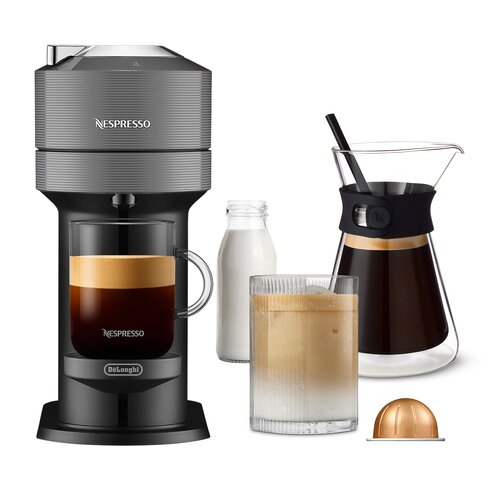 Na zdjęciu znajduje się szary ekspres do kawy z logo "Nespresso" na górnej części oraz "DeLonghi" na dolnej. Obok ekspresu widoczne są szklanka z kawą, butelka mleka, dzbanek z czarną opaską i słomką oraz złota kapsułka do kawy.
